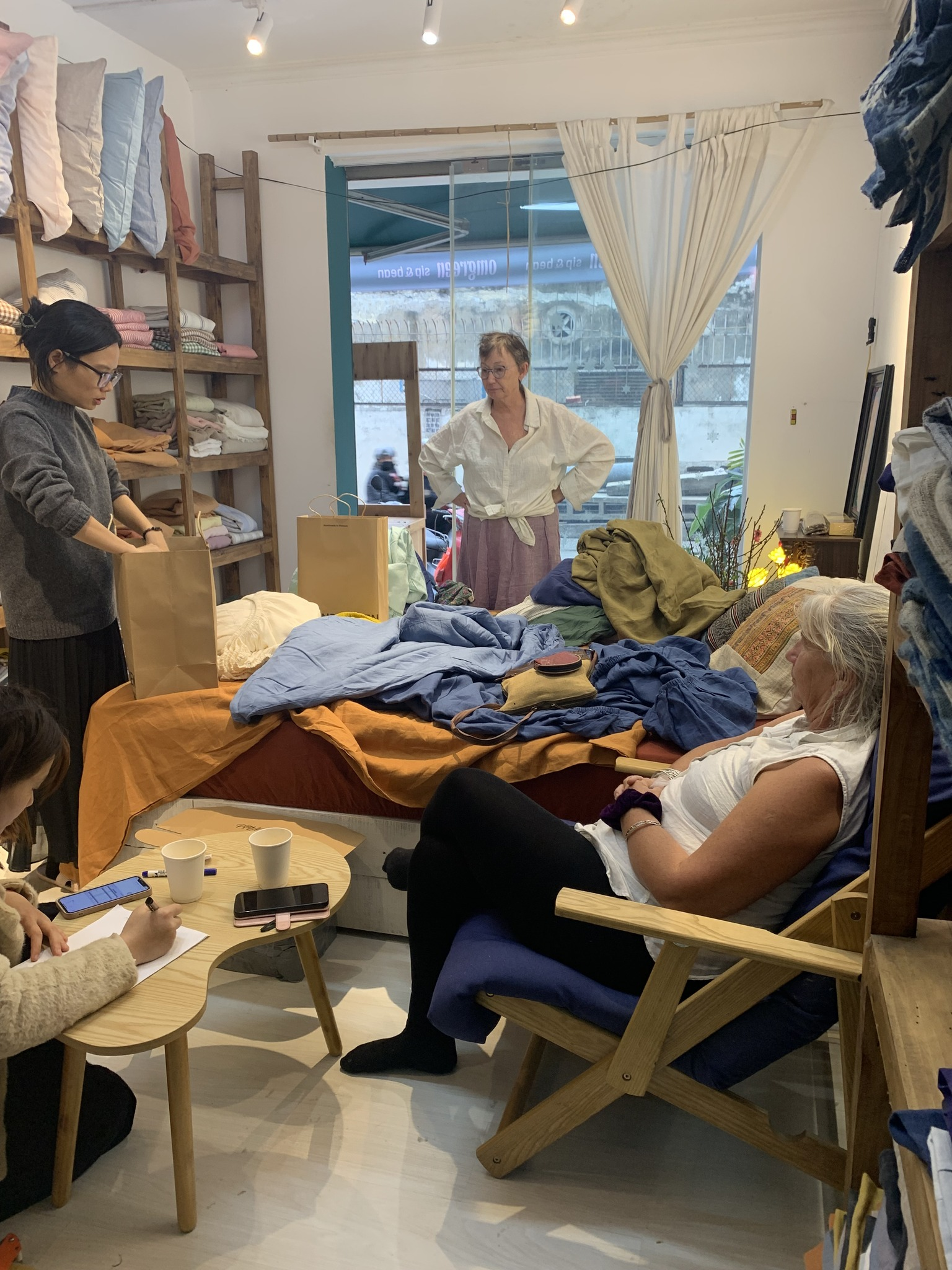 Inside the SCANDALINEN workshop in Hanoi — customers browsing French linen bedding in natural light with shelves of folded linen in the background