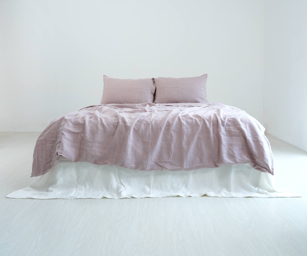 Dusty pink French linen duvet cover and matching pillows on a bed with a white bed skirt, shown in natural daylight.