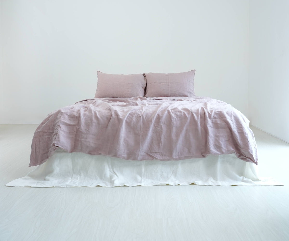 Dusty pink French linen duvet cover and matching pillows on a bed with a white bed skirt, shown in natural daylight.