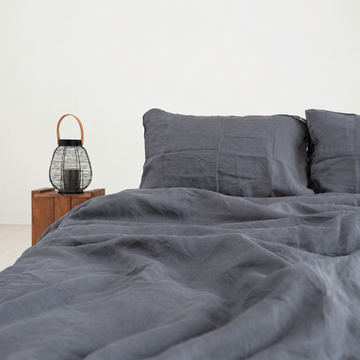Dark grey French linen duvet cover with natural folds, styled beside a wooden bedside table and wire lantern.
