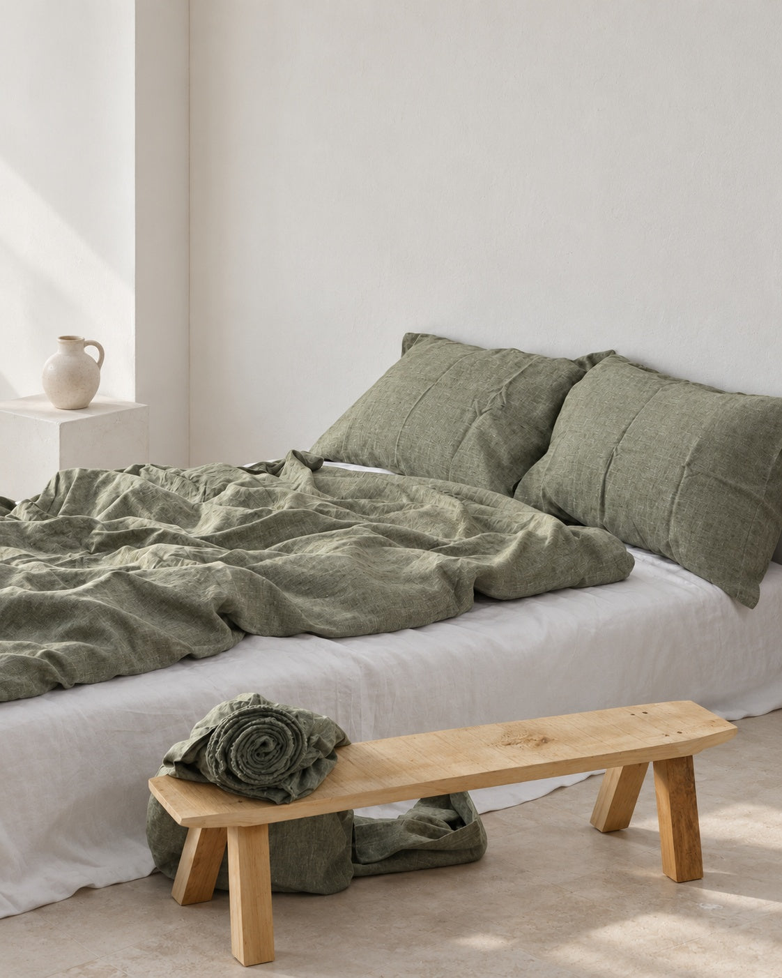 Asparagus olive green French linen duvet cover and matching pillows with a heathered weave, beside an oak bench in a white studio