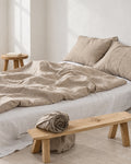 Natural flax French linen duvet cover and matching pillows with visible woven texture, styled beside an oak bench in a white studio