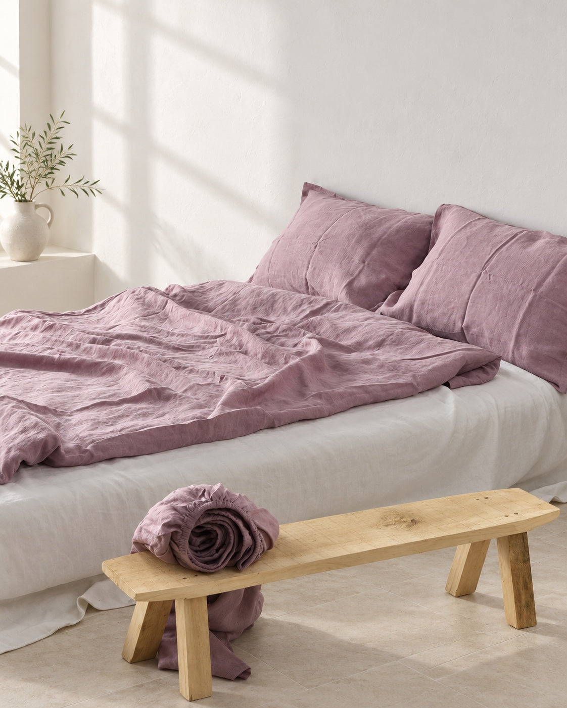 Mauve purple French linen duvet cover and matching pillows with natural fabric folds, beside an oak bench in a white studio